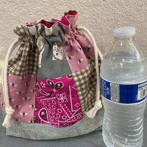 May include: A patchwork drawstring bag with various fabrics, including pink with bees, brown gingham, and a pink paisley pattern. The bag has a grey base and white drawstring cords. A water bottle is next to the bag.