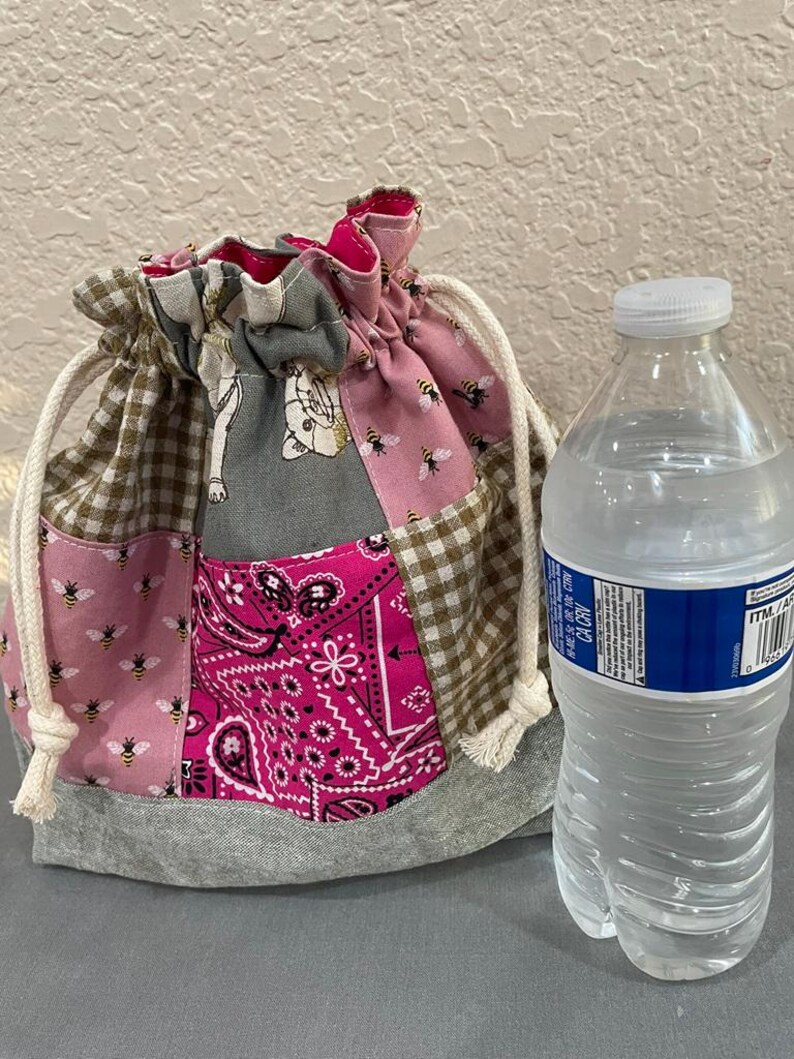 May include: A patchwork drawstring bag with various fabrics, including pink with bees, brown gingham, and a pink paisley pattern. The bag has a grey base and white drawstring cords. A water bottle is next to the bag.