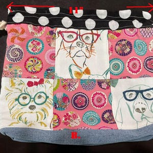 Pu&ograve; includere: Una borsa a coulisse patchwork, larga 28 cm e alta 23 cm, con illustrazioni di cani con gli occhiali. La borsa ha una base in denim, un motivo floreale rosa e una parte superiore a pois bianchi e neri.