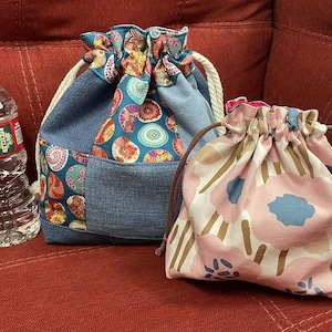 May include: Two fabric drawstring pouches. The larger pouch features blue denim panels and a colourful circular pattern with white rope cords. The smaller pouch has a pink, blue, and tan floral print with brown cords. A clear plastic water bottle is included.