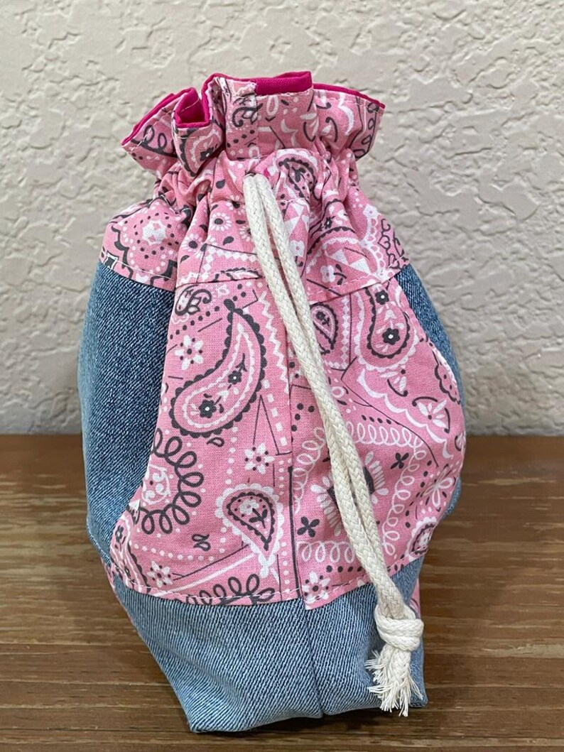 K&ouml;nnte beinhalten: Ein Kordelzugbeutel mit Jeans-Basis und einem rosa Paisley-Muster. Der Beutel hat einen wei&szlig;en Kordelzugverschluss und eine leuchtend rosa Einfassung. Der Beutel ist f&uuml;r kleine Gegenst&auml;nde konzipiert.