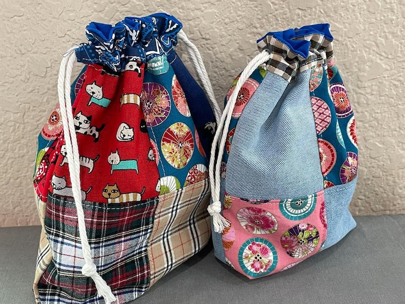 Może przedstawiać: Dwie torby ze sznurkiem ze wzorami patchwork. Jedna torba ma czerwoną tkaninę z ilustracjami kot&oacute;w, kratą i niebieskimi wzorami kwiatowymi. Druga torba ma dżins, r&oacute;żowe kwiaty i niebieskie tkaniny we wzory. Obie torby mają białe sznurki i niebieskie wzorzyste wierzchy.