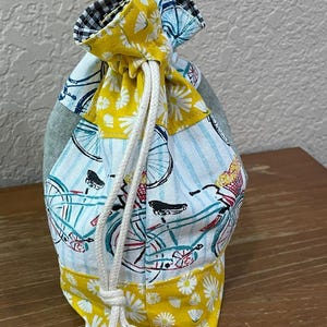 Op de afbeelding: Een trekkoordtas met een patchwork-ontwerp. De tas heeft een fietsprint op een lichtblauwe achtergrond, een gele madeliefjesprint en een zwart-wit geruite bovenkant. Een wit trekkoord wordt gebruikt om de tas te sluiten.