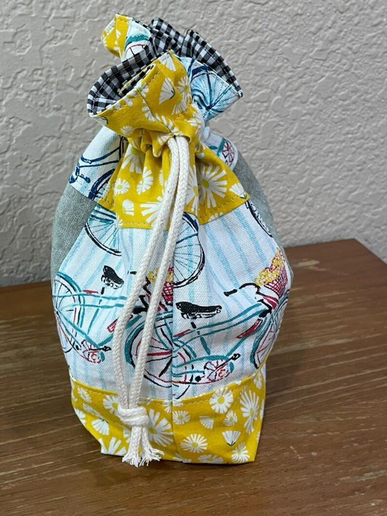 Op de afbeelding: Een trekkoordtas met een patchwork-ontwerp. De tas heeft een fietsprint op een lichtblauwe achtergrond, een gele madeliefjesprint en een zwart-wit geruite bovenkant. Een wit trekkoord wordt gebruikt om de tas te sluiten.