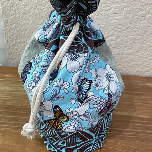 Op de afbeelding: Een trekkoordtas met een blauw, wit en bruin bloemen- en vlinderpatroon. De bovenkant is van donker denim. Een wit trekkoord is zichtbaar. De tas staat op een houten oppervlak.