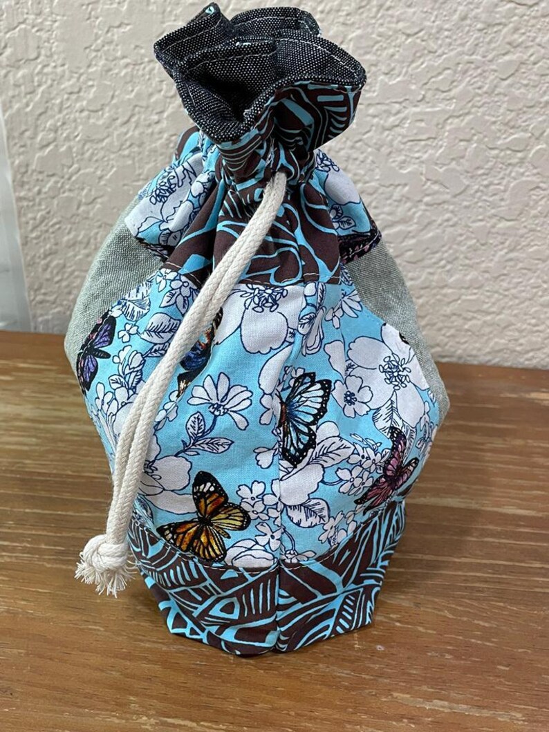 Op de afbeelding: Een trekkoordtas met een blauw, wit en bruin bloemen- en vlinderpatroon. De bovenkant is van donker denim. Een wit trekkoord is zichtbaar. De tas staat op een houten oppervlak.