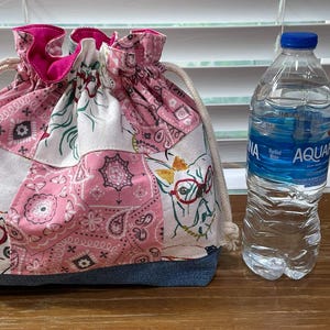 May include: A drawstring bag with a patchwork design in pink, white, and denim blue. The bag features a paisley pattern and a cat wearing glasses. A clear water bottle with a blue cap is next to the bag.