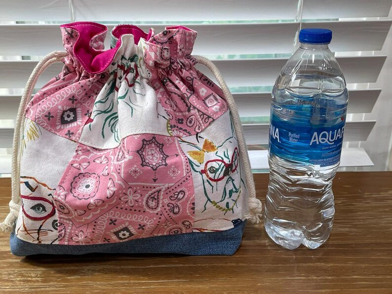 May include: A drawstring bag with a patchwork design in pink, white, and denim blue. The bag features a paisley pattern and a cat wearing glasses. A clear water bottle with a blue cap is next to the bag.