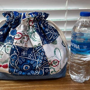 May include: A drawstring bag with a patchwork design featuring dogs wearing glasses and bow ties, and blue paisley patterns. The bag has a denim base and a cream-colored drawstring. A water bottle is next to the bag.