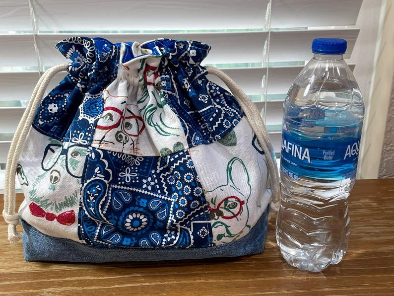 May include: A drawstring bag with a patchwork design featuring dogs wearing glasses and bow ties, and blue paisley patterns. The bag has a denim base and a cream-colored drawstring. A water bottle is next to the bag.