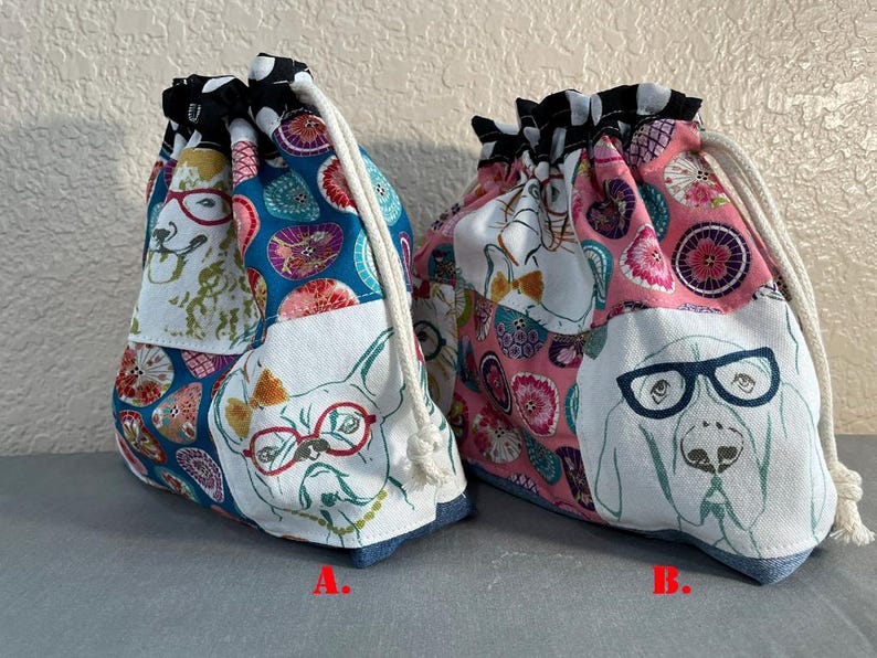 Pu&ograve; includere: Due sacchetti con coulisse con tessuto a tema cani. Una borsa presenta uno sfondo blu con illustrazioni di cani con gli occhiali, e l'altra ha uno sfondo rosa. Entrambe le borse hanno una parte superiore a fantasia bianca e nera e una base in denim.