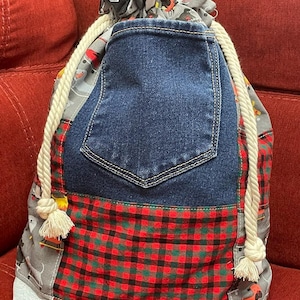 May include: A drawstring backpack constructed from a patchwork of fabrics, featuring a central dark blue denim pocket, a red and black buffalo plaid section, and a grey fabric printed with various dog illustrations. It has thick white rope straps.