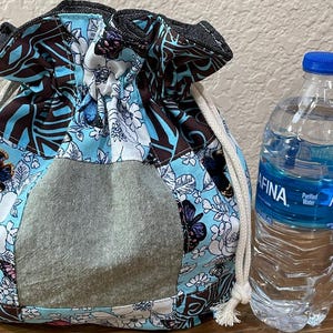 Op de afbeelding: Een trekkoordtas met een bloemen- en vlinderpatroon in tinten blauw, bruin en wit. De tas heeft een grijze zak en een wit trekkoord. Naast de tas staat een doorzichtige plastic waterfles met een blauwe dop.