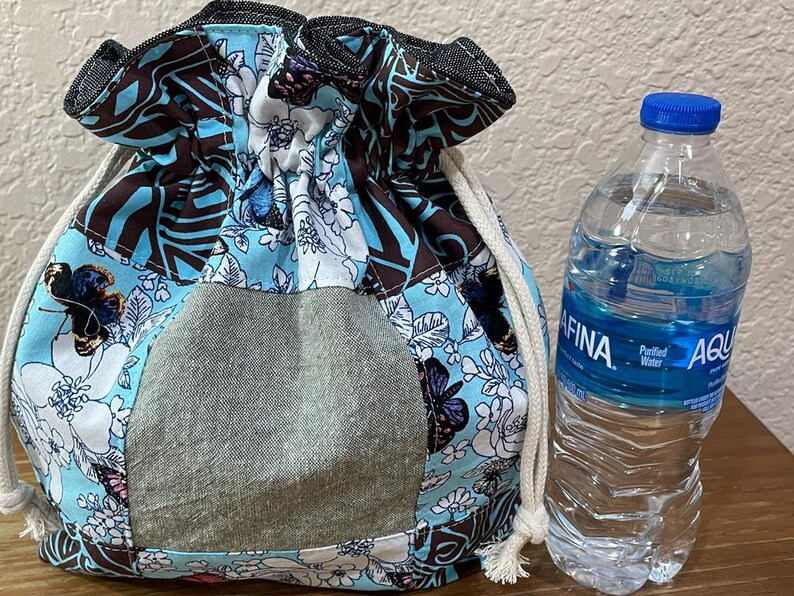 Op de afbeelding: Een trekkoordtas met een bloemen- en vlinderpatroon in tinten blauw, bruin en wit. De tas heeft een grijze zak en een wit trekkoord. Naast de tas staat een doorzichtige plastic waterfles met een blauwe dop.