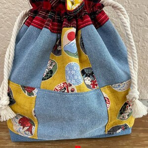 May include: A drawstring bag with a patchwork design. The bag features denim, yellow fabric with Japanese-style illustrations, and red plaid fabric at the top. White rope handles with tassels complete the design.