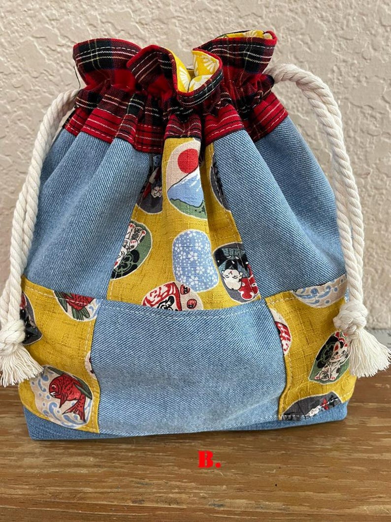 May include: A drawstring bag with a patchwork design. The bag features denim, yellow fabric with Japanese-style illustrations, and red plaid fabric at the top. White rope handles with tassels complete the design.
