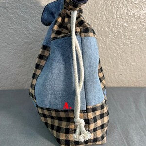 May include: A drawstring bag with a patchwork design. The bag features denim blue fabric, tan and black checkered fabric, and a navy blue fabric at the top. A white drawstring is used to close the bag.