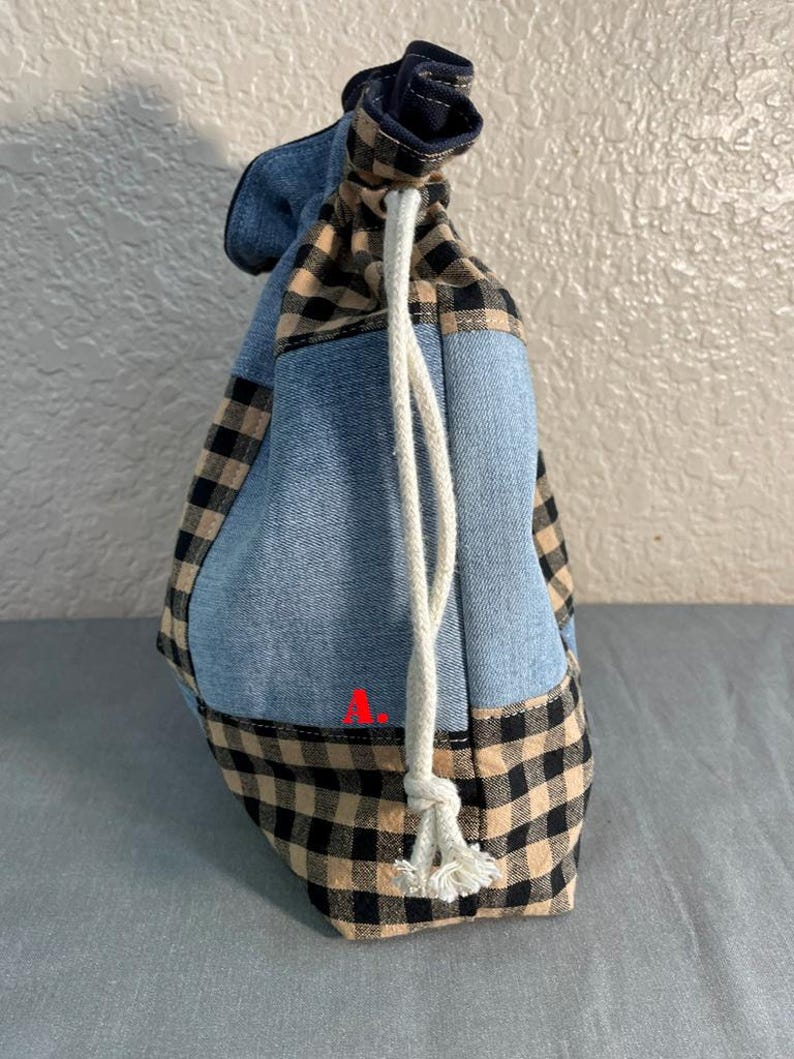 May include: A drawstring bag with a patchwork design. The bag features denim blue fabric, tan and black checkered fabric, and a navy blue fabric at the top. A white drawstring is used to close the bag.