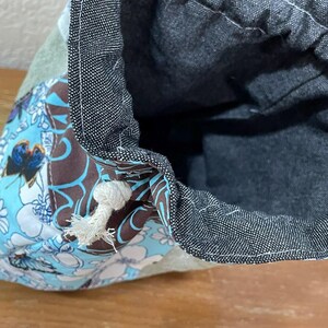 Op de afbeelding: Close-up van een trekkoordtas met een bloemenpatroon in blauw, bruin en wit. De binnenkant van de tas is donkerblauw en de bovenrand is afgewerkt met een donkere denim stof. Een wit trekkoord is zichtbaar.