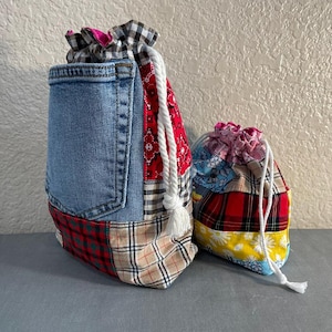 May include: Two drawstring bags. The larger bag features a denim pocket and patchwork of plaid and patterned fabrics. The smaller bag has a patchwork design with red, yellow, and blue fabrics. Both bags have white drawstring cords.