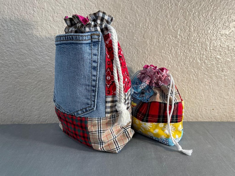May include: Two drawstring bags. The larger bag features a denim pocket and patchwork of plaid and patterned fabrics. The smaller bag has a patchwork design with red, yellow, and blue fabrics. Both bags have white drawstring cords.