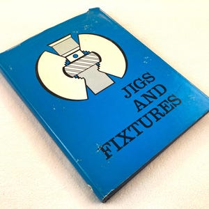 May include: A blue hardcover book titled JIGS AND FIXTURES in black capital letters. The cover features a graphic illustration of a mechanical chuck component in white, gray, and black, positioned above the title text on the front cover.