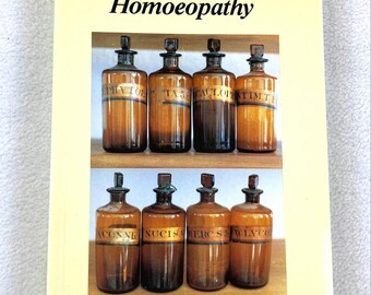 May include: A softcover book titled Talking About Homoeopathy by Dr. Trevor Smith. The cover is pale yellow with black text and a central rectangular image showing eight amber glass apothecary bottles with stoppers arranged on two wooden shelves.