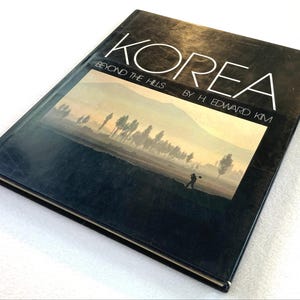 May include: A hardcover book titled KOREA BEYOND THE HILLS BY H. EDWARD KIM. The black cover features a rectangular photograph of a hazy landscape with silhouetted trees and a person walking in a field under a pale yellow sky.