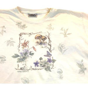 May include: Cream-colored t-shirt with a nature-themed design. The shirt features a central illustration of mushrooms, flowers, and a butterfly within a vine frame. Additional floral and botanical sketches are scattered across the fabric.