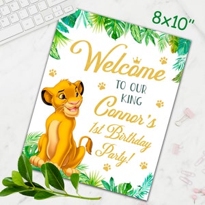 May include: A welcome sign for a first birthday party with a jungle theme. The sign features a cartoon lion cub, green leaves, and gold lettering that says "Welcome to our King Connor's 1st Birthday Party!"