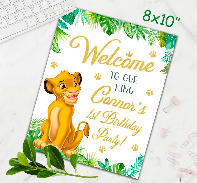 May include: A welcome sign for a first birthday party with a jungle theme. The sign features a cartoon lion cub, green leaves, and gold lettering that says "Welcome to our King Connor's 1st Birthday Party!"