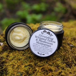 Puede incluir: Dos pequeños frascos de vidrio con tapas negras llenos de una sustancia amarilla similar a la crema batida. El frasco de la derecha tiene una etiqueta blanca con texto negro que dice "Grounding Whipped Tallow & Mushroom Moisturizer All-Natural Small Batch - 2 oz Ingredients: Grass-fed beef tallow, grass-fed shea butter, jojoba oil, cocoa butter, reishi & chaga mushroom extract, Melrose & frankincense essential oils."
