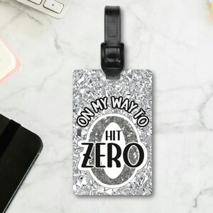May include: A rectangular luggage tag with a black leather strap. The tag has a silver glitter background with the words "ON MY WAY TO HIT ZERO" in black and white. A keyboard, notebook, and pens are in the background.