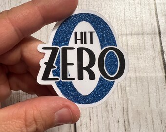 Peut inclure: Un autocollant bleu et blanc avec le texte "HIT ZERO" en lettres noires. L'autocollant a une texture scintillante.