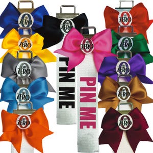 May include: A collection of cheer bows in various colors, each featuring a central circular emblem with the text HIT ZERO. Two white ribbons hang vertically from the center, each printed with the text PIN ME in black or pink ink.