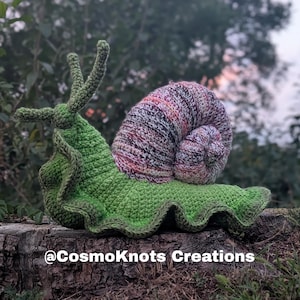 May include: A crocheted snail toy featuring a bright green body with a ruffled edge and a spiraled shell in variegated shades of purple, pink, white, and black. The text @CosmoKnots Creations is printed in white at the bottom of the image.