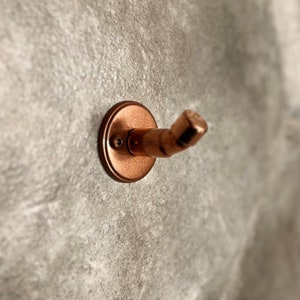 May include: A copper-colored wall hook with a rounded base and a curved arm. The hook is mounted on a textured gray wall.