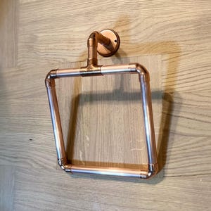 Pode incluir: Um porta-toalhas de cobre com forma quadrada e base redonda. O anel está montado em uma parede.
