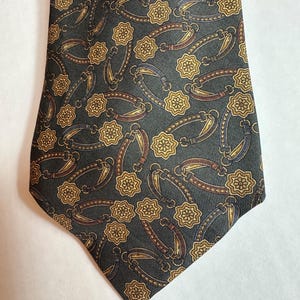 Puede incluir: Una corbata gris oscuro con un patrón repetido de formas florales doradas y marrones y diseños ovalados. Los diseños ovalados tienen acentos marrones, dorados y azules. La corbata está hecha de una tela suave.