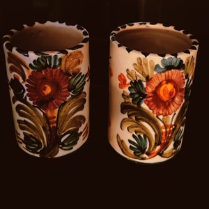 May include: Two white ceramic cups with floral designs. The cups have green, red, orange, and yellow flowers and leaves. The cups are perfect for holding pens, pencils, or other small items.