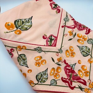 May include: A light peach-colored fabric featuring a repeating pattern of orange fruits, green leaves, and red floral accents. The textile is divided by thin green lines and finished with a distinct red border, creating a structured geometric layout.