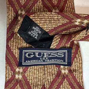 May include: A patterned silk tie with a diamond design in shades of brown and burgundy. The tie features a label that reads "GUESS U.S.A. AMERICAN TRADITION" and a tag that says "100% Imported Silk Made in U.S.A."