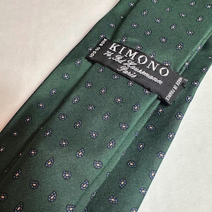 May include: A dark green silk tie with a repeating pattern of small blue and tan floral designs. The label reads "KIMONO 74, Bd Haussmann Paris" and "Made in France". The tie is 100% silk.