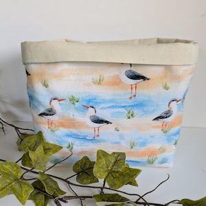May include: A fabric storage basket with a rolled-down top. The exterior features a watercolour design of seagulls on a beach with blue water, tan sand, and green grass. The top and interior are a solid beige colour.