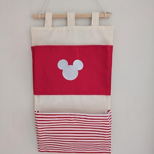 May include: A hanging wall organiser made of off-white fabric with a wooden dowel and string. It features two pockets: the top pocket is solid red with a white mouse head silhouette, and the bottom pocket has a red and white horizontal stripe pattern.