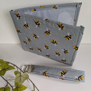 May include: A grey fabric wallet and matching wrist strap, both patterned with yellow and black bumblebees. The wallet is open, showing a light interior. The wrist strap has a silver metal ring.