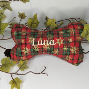 May include: A dog bone-shaped toy in red and green plaid, decorated with gold snowflakes. The name "Luna" is written on the toy. The toy is surrounded by green leaves and brown vines.
