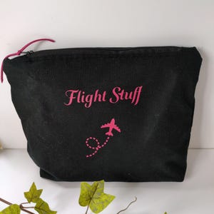 May include: Black travel pouch with the words "Flight Stuff" in pink script. A pink airplane graphic with a dotted trail is below the text. The pouch has a pink zip pull and is likely made of canvas or a similar fabric.