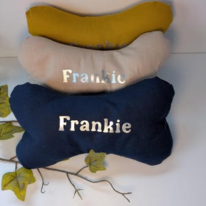 May include: Three dog bone-shaped cushions in mustard yellow, beige, and navy blue, stacked. Each cushion has the name "Frankie" printed in silver. A decorative item for pets.
