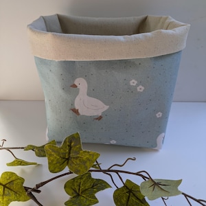 May include: A fabric storage basket with a pale blue background, patterned with white ducks and small white flowers. The top edge and interior lining are a natural beige colour. A decorative vine with green leaves is in the foreground.